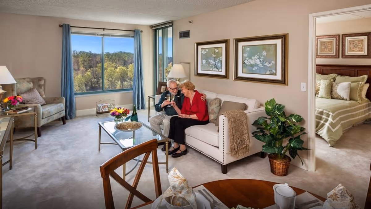A cozy living room with a large window showing a view of trees outside. An elderly couple sits on a white couch, smiling and holding glasses. The room has a glass coffee table with flowers and a decorative bowl, an armchair with a pillow, and framed floral artwork on the wall. To the right, an open doorway reveals a bedroom with a neatly made bed and decorative pillows. A potted plant is placed near the doorway.