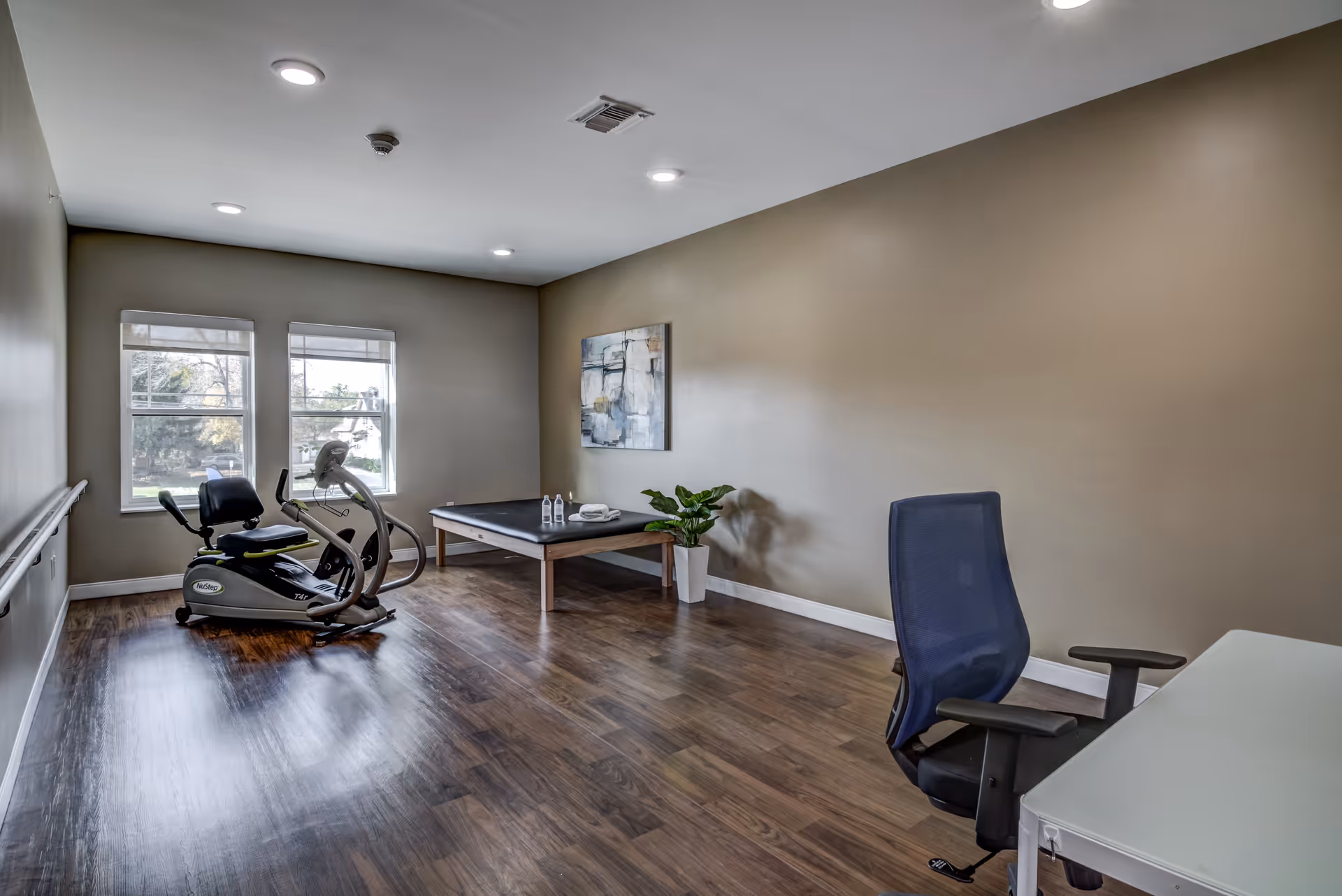 A room with wooden flooring and beige walls featuring a recumbent exercise bike near two windows, a black padded therapy table with two water bottles and a towel on it, a potted plant, an abstract painting on the wall, and a black office chair next to a white desk.