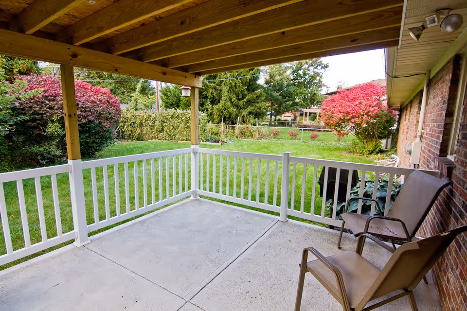 Covered patio area with a wooden roof and white railing overlooking a green lawn with bushes and trees. Two brown chairs are placed on the concrete floor near a brick wall of the building.