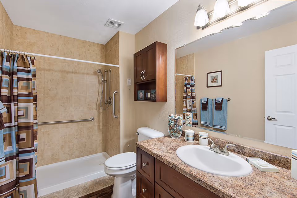 Well-lit bathroom with a sink vanity, toilet, and a tiled shower with a patterned curtain and grab bars.