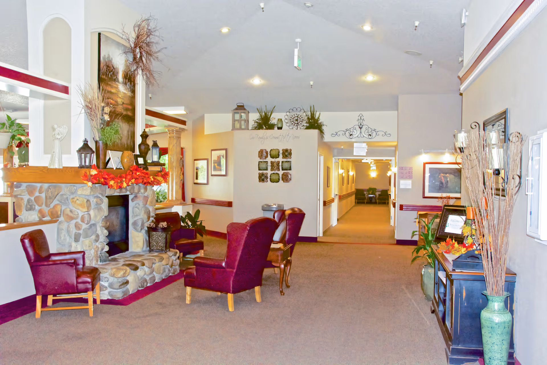 Interior view of a senior living facility lounge area with a stone fireplace decorated with autumn leaves and various vases. There are several burgundy armchairs arranged around the fireplace. The hallway extends into the background with framed artwork on the walls and decorative plants placed throughout the space.