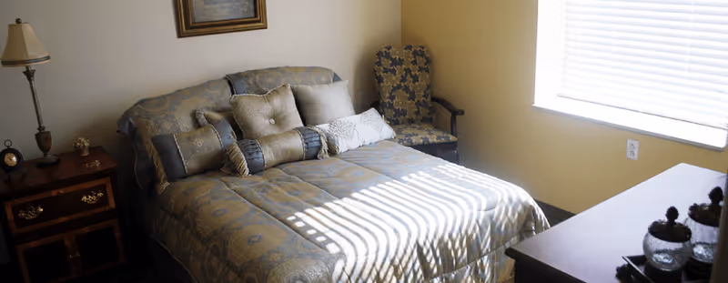 A bedroom with a neatly made bed featuring multiple decorative pillows and a patterned bedspread. To the left of the bed is a wooden nightstand with a lamp and a small clock. In the corner near the window, there is a cushioned armchair with a floral pattern. Sunlight streams through the window blinds, casting shadows on the bed.
