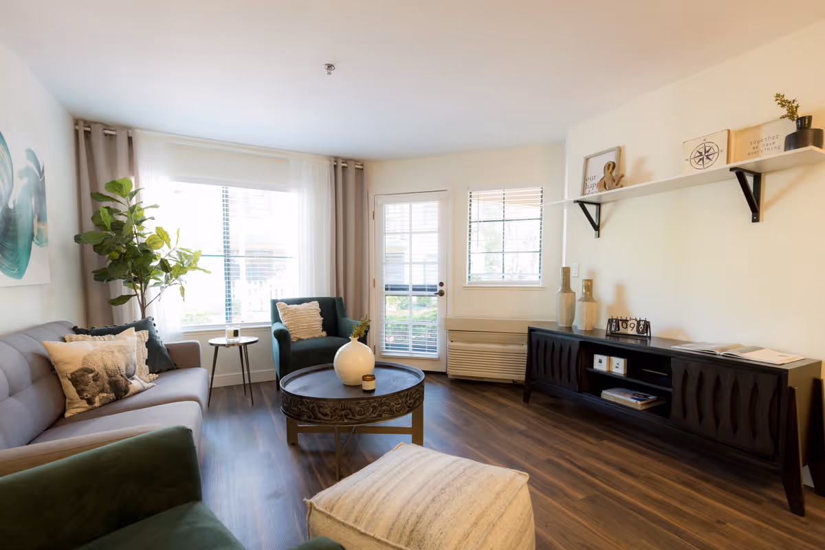A bright living room with a gray sofa, green armchairs, a round coffee table with a white vase and candle, a small side table, a large window with curtains, a door with glass panels, a window with blinds, a dark wooden media console with decorative items, and a white floating shelf with framed pictures and a small plant.