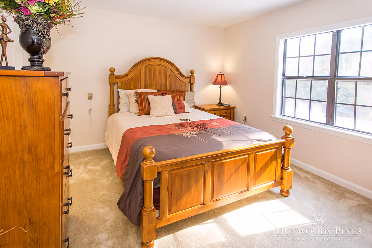 A bright bedroom with a wooden bed frame and headboard, a bed made with a multicolored comforter and several pillows, a wooden dresser with a decorative vase of flowers on top, a wooden nightstand with a lamp, and a large window letting in natural light.
