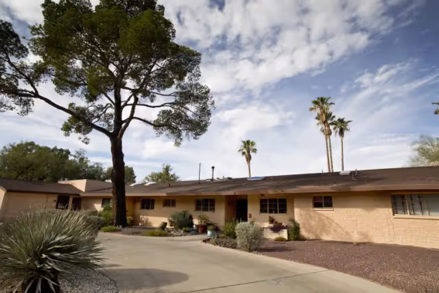 Single-story beige building with a flat roof surrounded by desert landscaping including bushes, a large tree, and palm trees under a partly cloudy sky.
