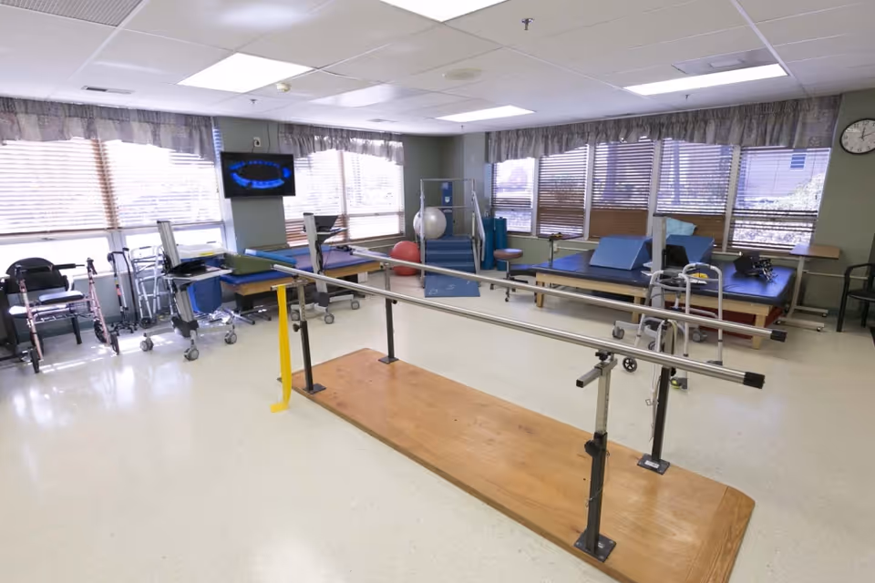 A rehabilitation therapy room with parallel bars for walking exercises in the center, exercise mats and therapy equipment along the walls, several walkers and wheelchairs near the windows, and a wall-mounted TV. The room has large windows with blinds and curtains, and a clock on the wall.