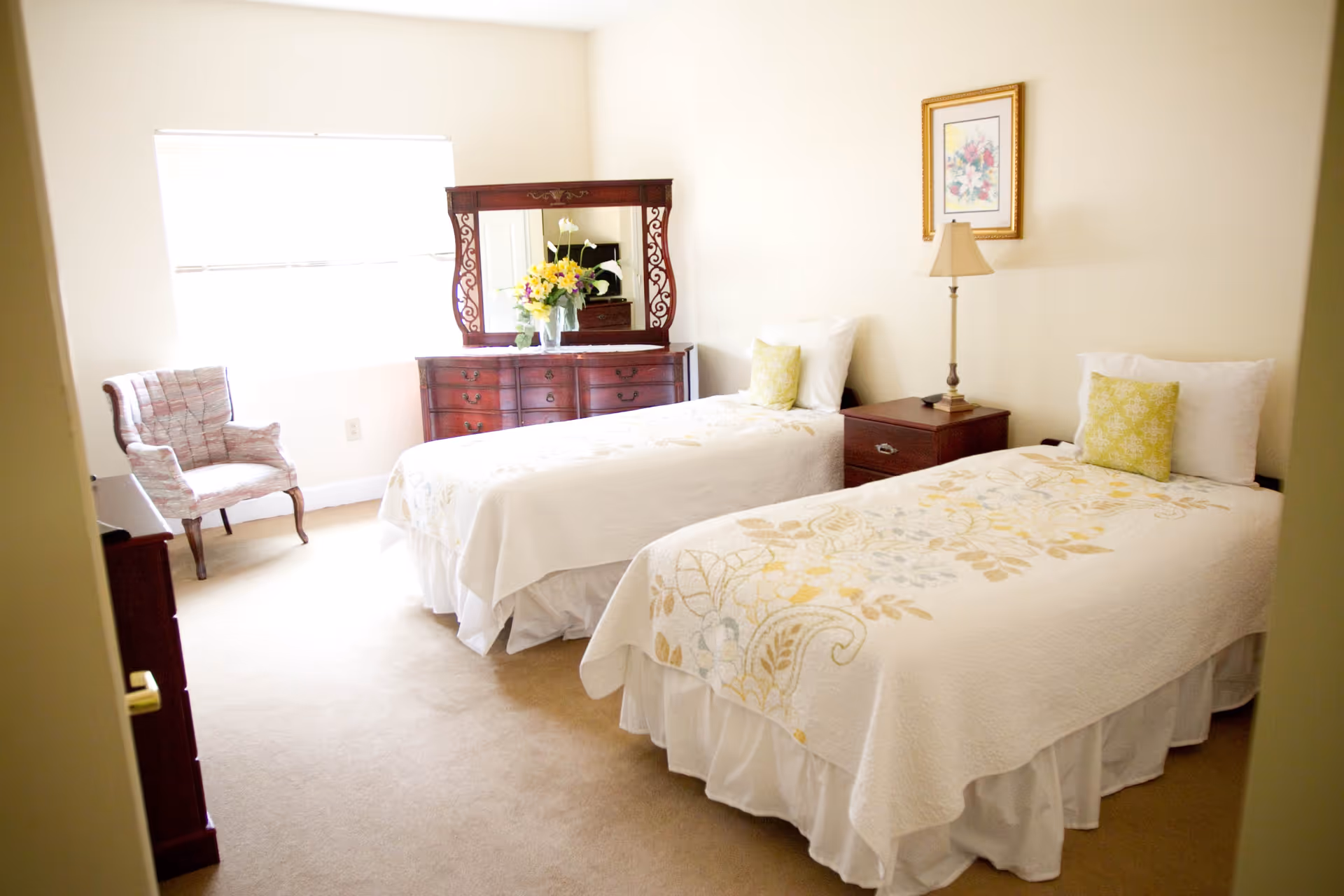 A bright bedroom with two twin beds covered in white and floral bedspreads. Each bed has a white pillow and a decorative yellow pillow. Between the beds is a wooden nightstand with a lamp and a framed floral picture hanging above. Across from the beds is a wooden dresser with an ornate mirror and a vase of yellow and white flowers. A cushioned armchair is placed near a window with blinds, allowing natural light to fill the room.