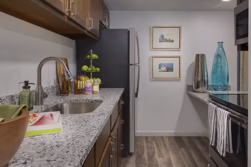 Modern kitchen with granite countertops, stainless steel sink and faucet, wooden cabinets, a stainless steel refrigerator, and stove. Decorative items including a tiered fruit stand with green apples, bottles, and vases are placed on the counters. Two framed pictures hang on the white wall at the end of the kitchen.