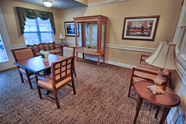 A cozy interior room with a wooden table surrounded by four chairs, a cushioned bench under a window with green curtains, a wooden display cabinet against the wall, framed artwork, and a side table with a lamp.