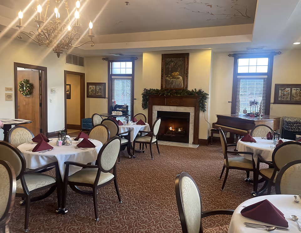 A cozy dining room with round tables covered in white tablecloths and set with burgundy napkins. The room features a lit fireplace with a decorative garland and a painting above it. There are two windows with blinds, a chandelier hanging from the ceiling, and a piano in the corner. The carpet has a patterned design, and the walls are adorned with framed artwork.