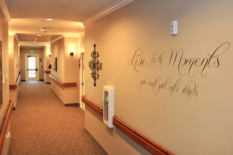 A carpeted, well-lit interior hallway with wooden handrails, wall sconces, decorative wall art and a script reading 'Live for the Moments'.
