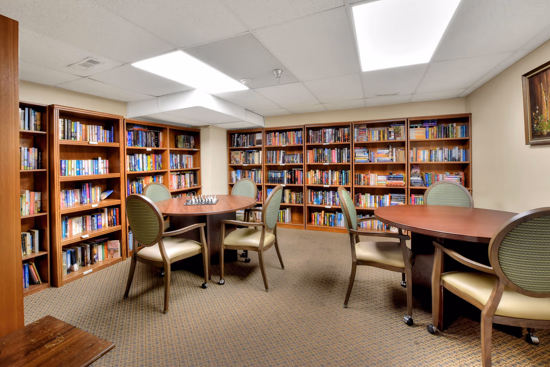 A cozy library room with multiple wooden bookshelves filled with books lining the walls. There are two round wooden tables, each surrounded by four cushioned chairs with green backs and beige seats. A chessboard is set up on one of the tables. The room has a carpeted floor and a drop ceiling with fluorescent lighting. A framed painting hangs on the right wall.