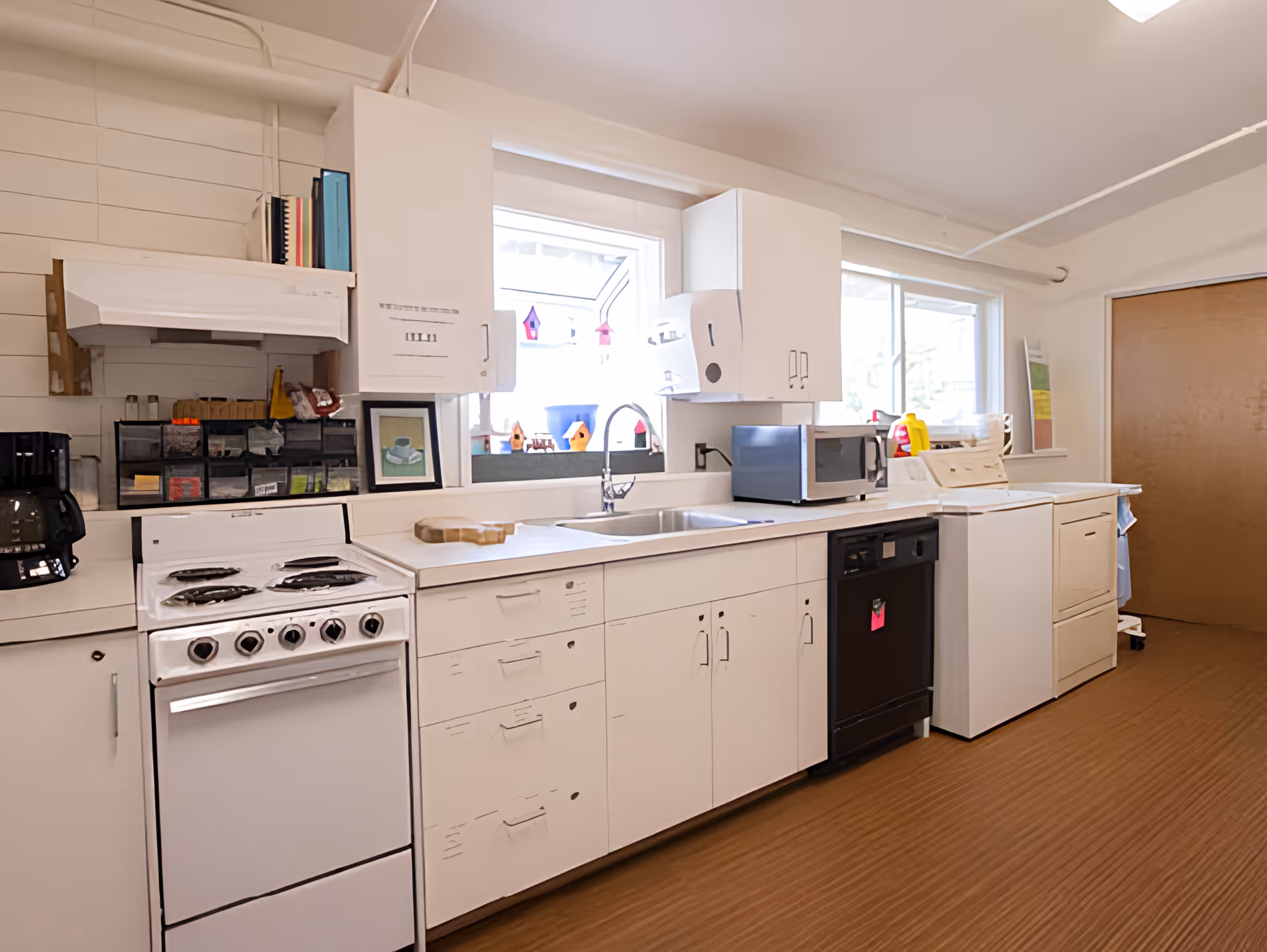 A clean kitchen area with white cabinets and countertops, featuring a stove with four burners, a coffee maker, a microwave, a dishwasher, a washing machine, and a dryer. There are two windows above the sink with small decorative birdhouses on the windowsill. The floor has a wood-like finish and there is a closed wooden door at the far end of the room.