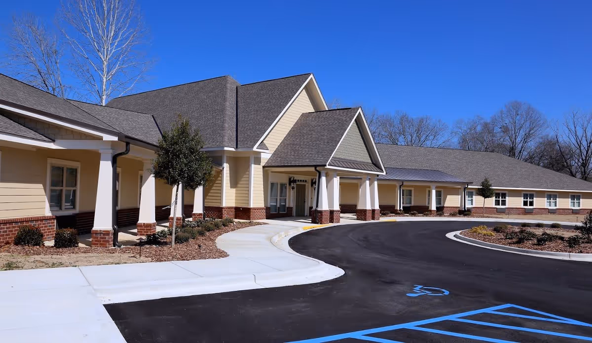 Exterior view of a single-story building with beige siding and brick accents, featuring a covered entrance with white columns and a curved driveway with a handicap parking space marked in blue. Leafless trees and clear blue sky are visible in the background.