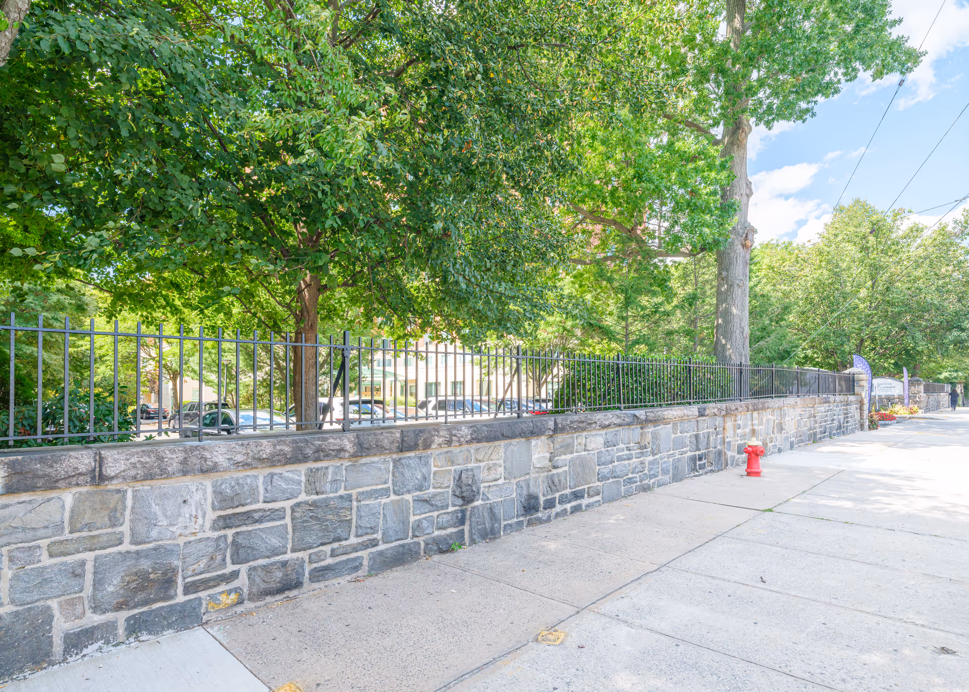 Stone wall topped with an iron fence along a tree-lined sidewalk and a red fire hydrant in front of the building grounds.