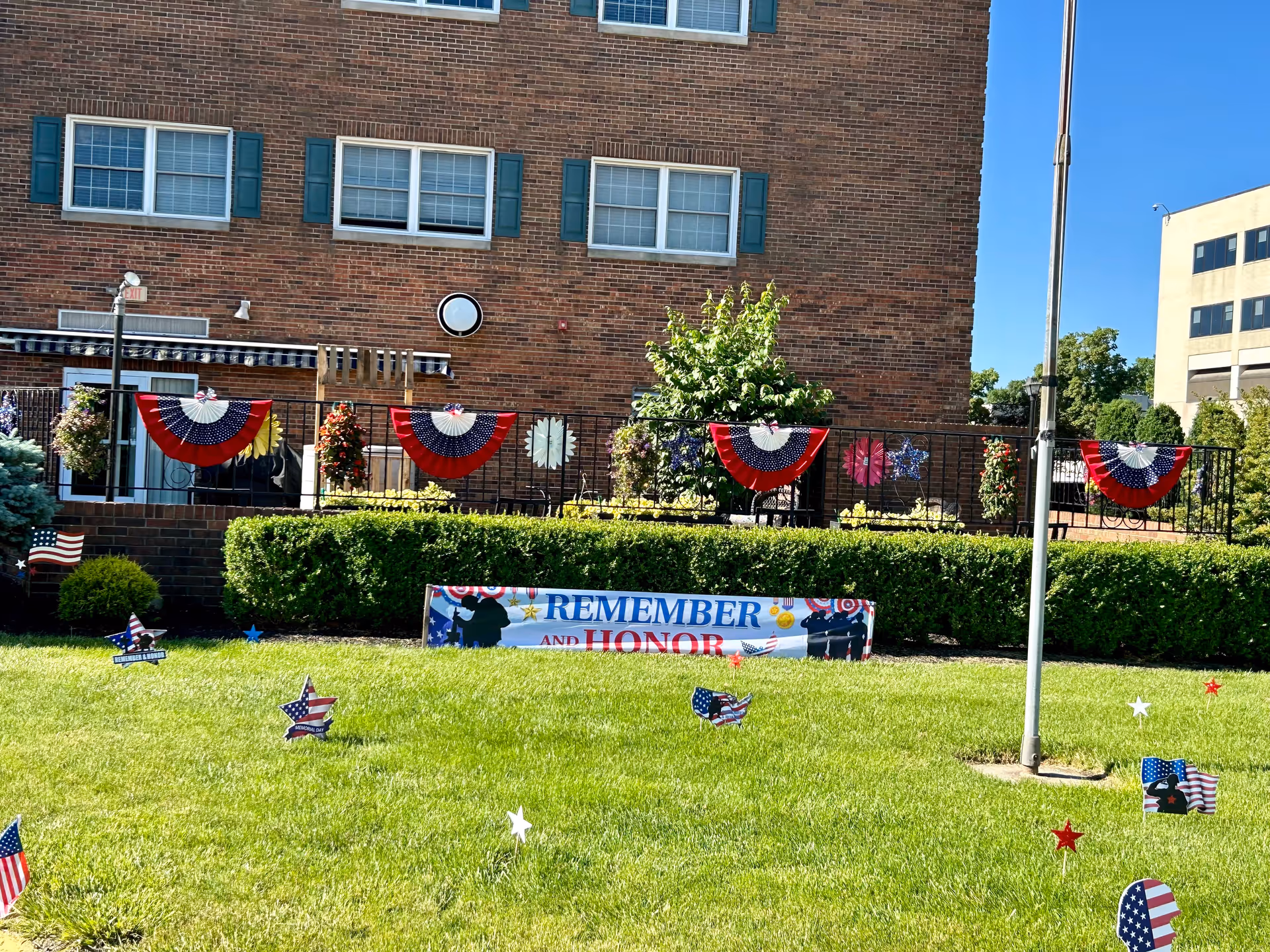 Outdoor view of a brick building with a decorated patio area featuring red, white, and blue patriotic bunting. The lawn in front has small American flag-themed decorations and a banner that reads 'REMEMBER AND HONOR'. A flagpole is visible on the right side of the image under a clear blue sky.