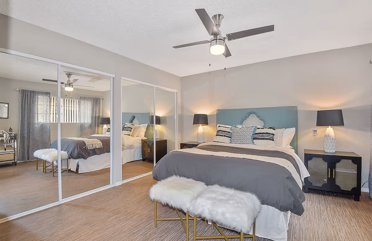 A well-lit bedroom featuring a bed with a teal upholstered headboard, gray and white bedding, and decorative pillows. On either side of the bed are black nightstands with white lamps. A bench with white furry cushions and gold legs is placed at the foot of the bed. The room has light wood flooring and mirrored sliding closet doors reflecting the bed and window with gray curtains.