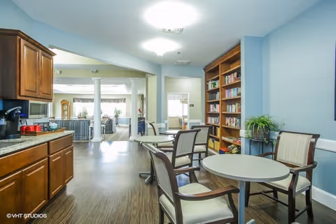 Interior view of a senior living facility common area with round tables and chairs, wooden cabinets and countertop on the left, a bookshelf filled with books on the right, and a seating area with couches and large windows in the background.