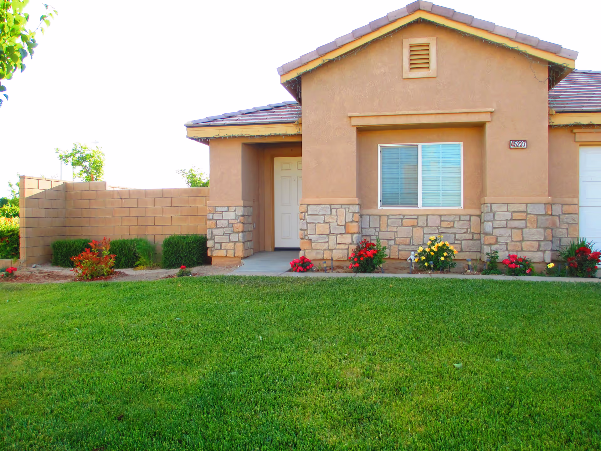 Front exterior view of a single-story house with a beige stucco and stone facade, a white front door, a window with closed blinds, a small porch, and a well-maintained green lawn with colorful flowers along the walkway.