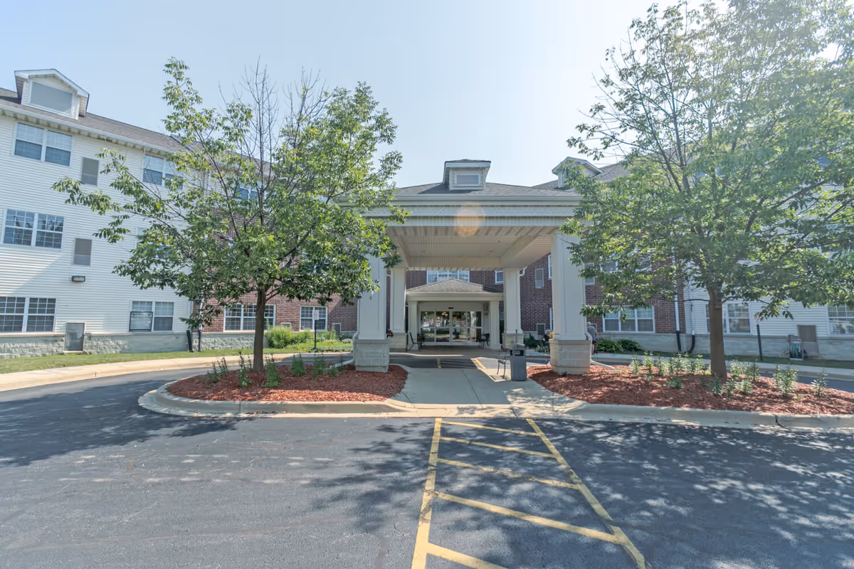 Front entrance of a senior living facility with a covered drop-off area, surrounded by trees and landscaping with red mulch. The building has white siding and brick exterior walls with multiple windows.