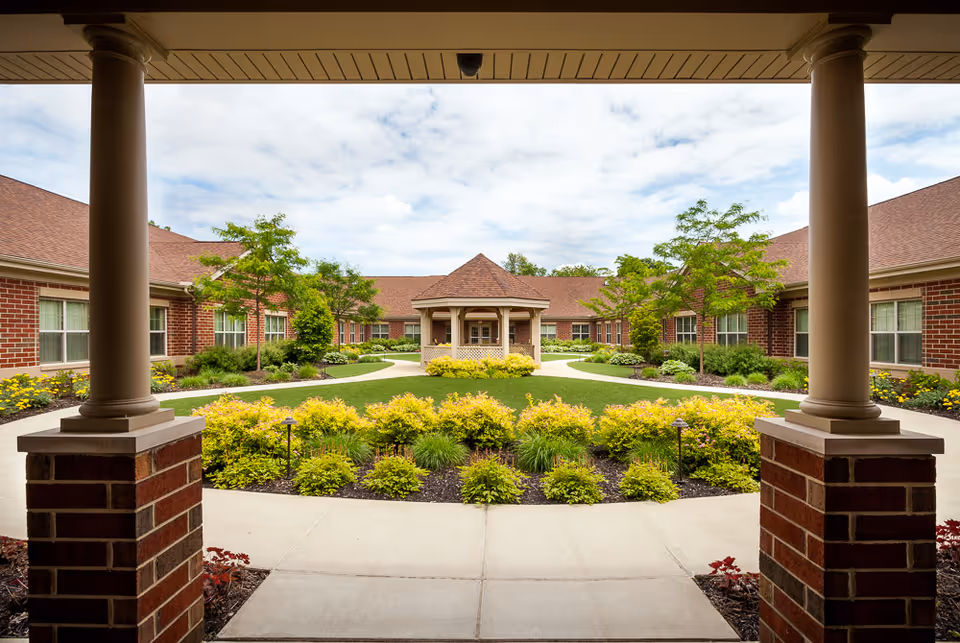 View of a landscaped courtyard with a gazebo in the center, surrounded by a brick building with multiple windows. The courtyard features green grass, yellow flowering bushes, and small trees, with a paved walkway encircling the garden area. The photo is taken from under a covered porch supported by two brick columns.