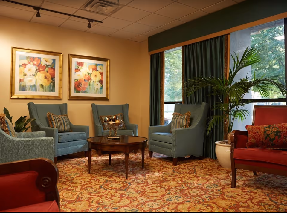 Well-furnished sitting area with upholstered armchairs around a coffee table, framed floral artwork, patterned carpet and large windows with plants.
