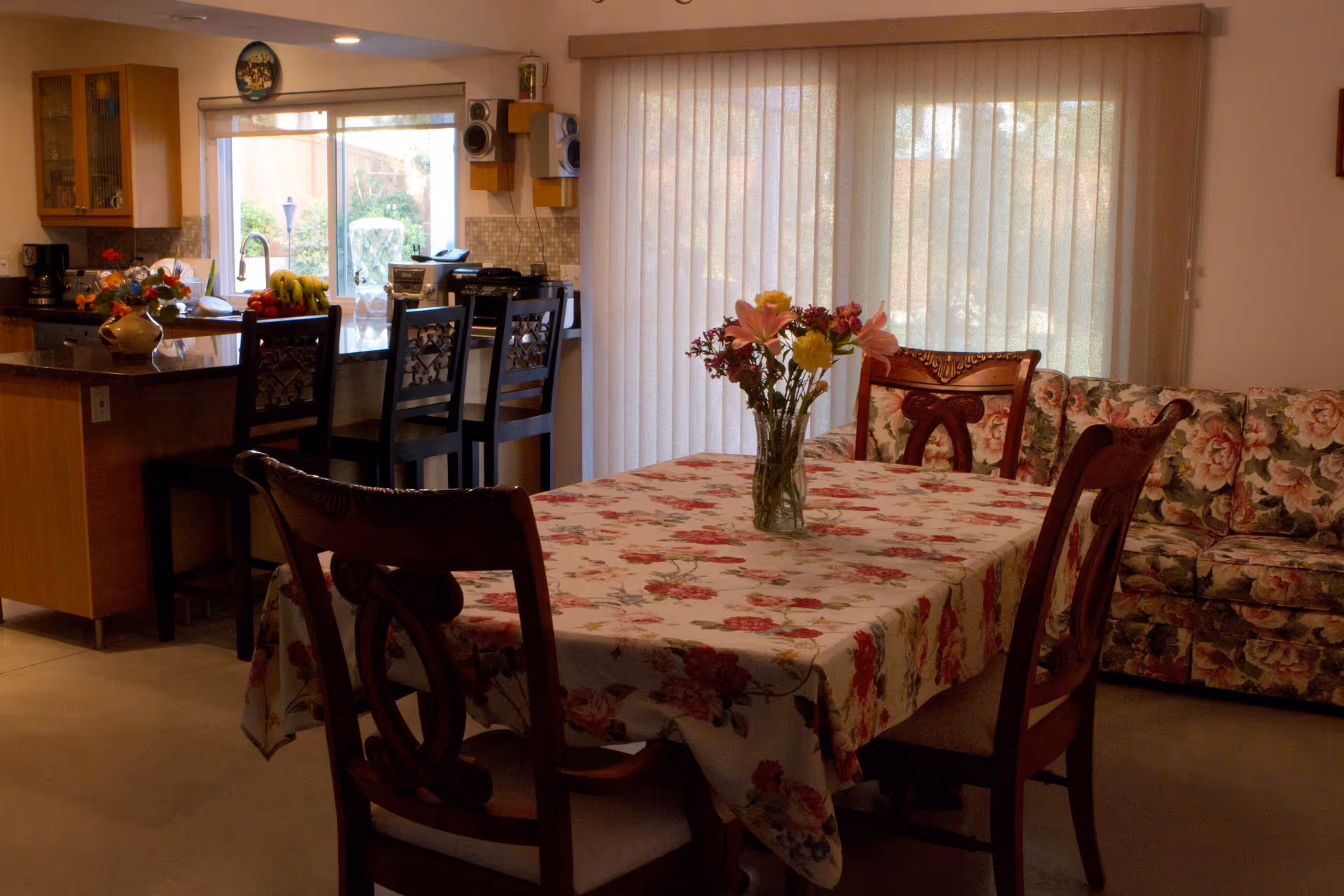 Open dining area with a floral tablecloth and vase of flowers, wooden chairs, a floral sofa, and a kitchen island in the background.