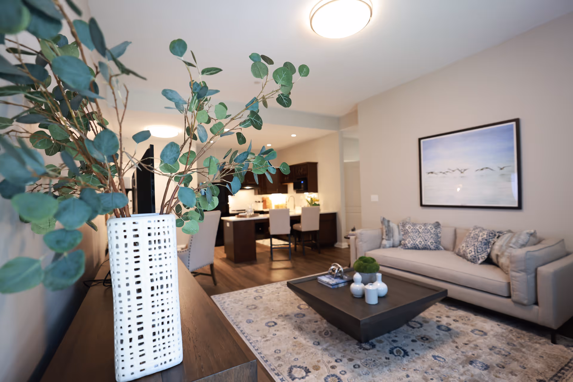 A modern living room with a beige sofa adorned with patterned pillows, a dark wooden coffee table with decorative items, and a large framed artwork of birds flying over water on the wall. In the foreground, there is a white vase with green leafy branches on a wooden console table. The room has wooden flooring and a soft patterned area rug. In the background, a kitchen with dark cabinets and a dining area with chairs is visible.