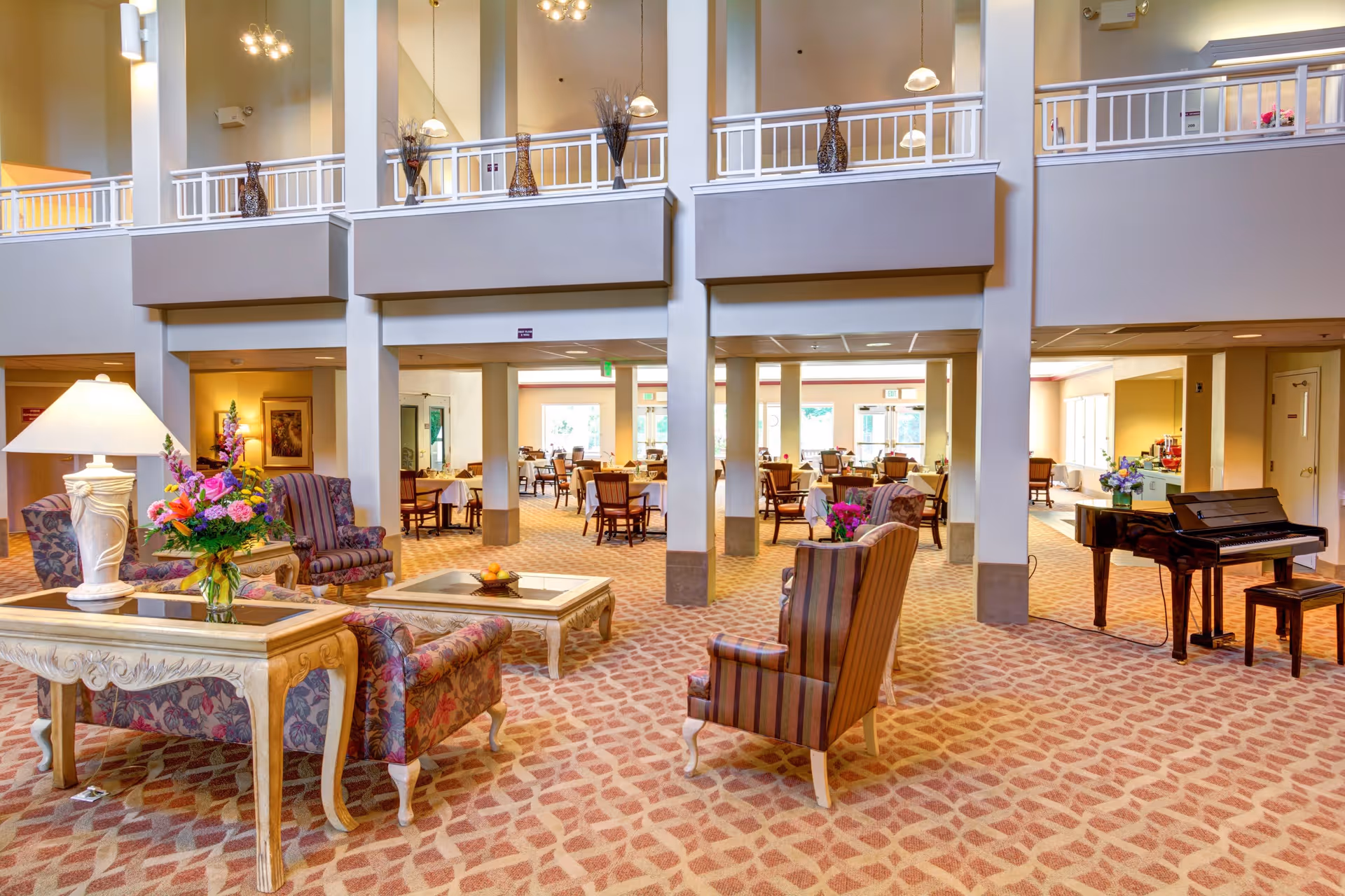 Spacious senior living facility common area with patterned carpet, multiple armchairs and tables with floral arrangements, a grand piano, and a dining area with tables and chairs in the background. The room has high ceilings with a balcony railing above and warm lighting.