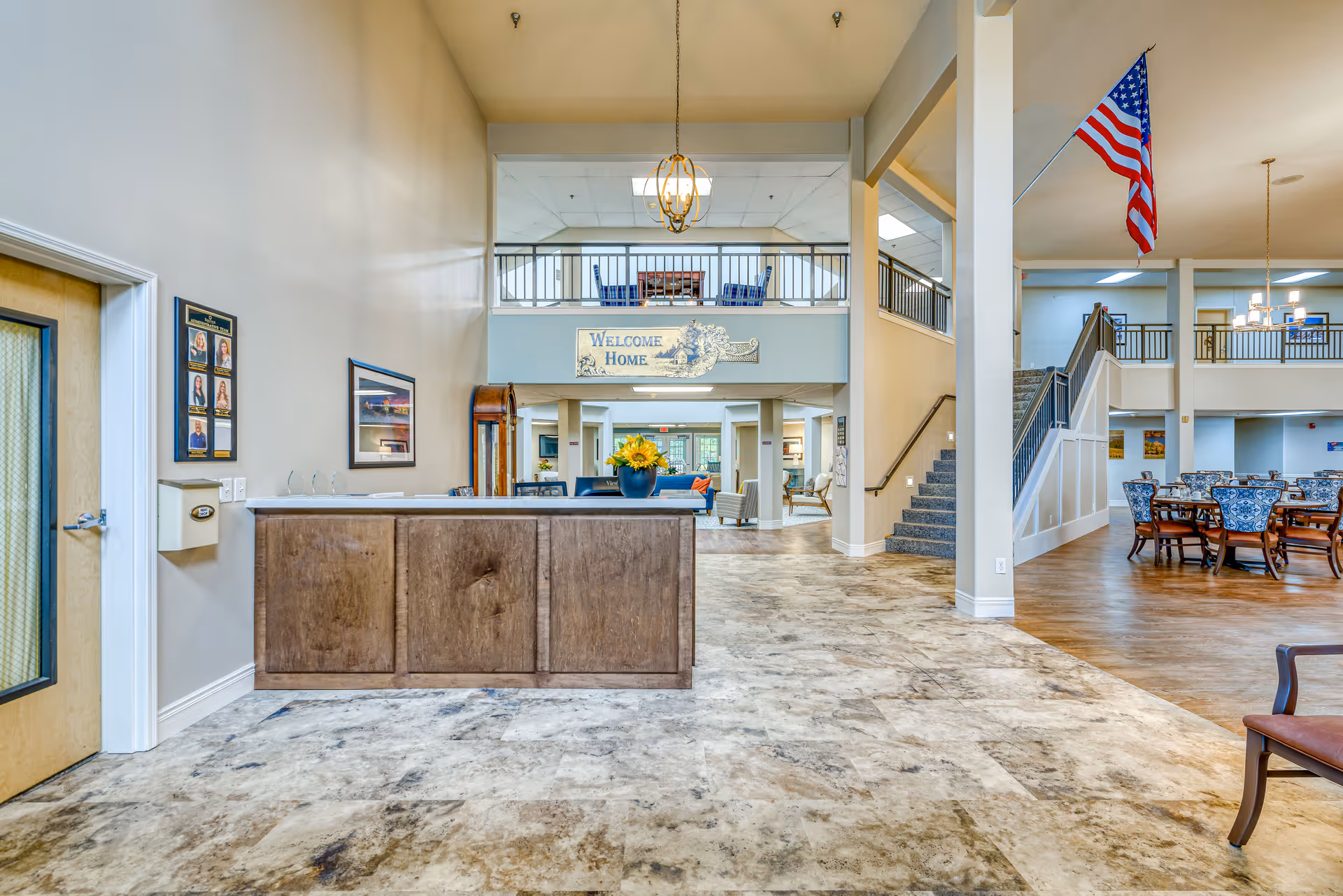 Interior view of a senior living facility lobby with a reception desk in the foreground, a 'Welcome Home' sign above a hallway, seating areas with chairs and tables, a staircase leading to an upper level, and an American flag hanging from the ceiling.