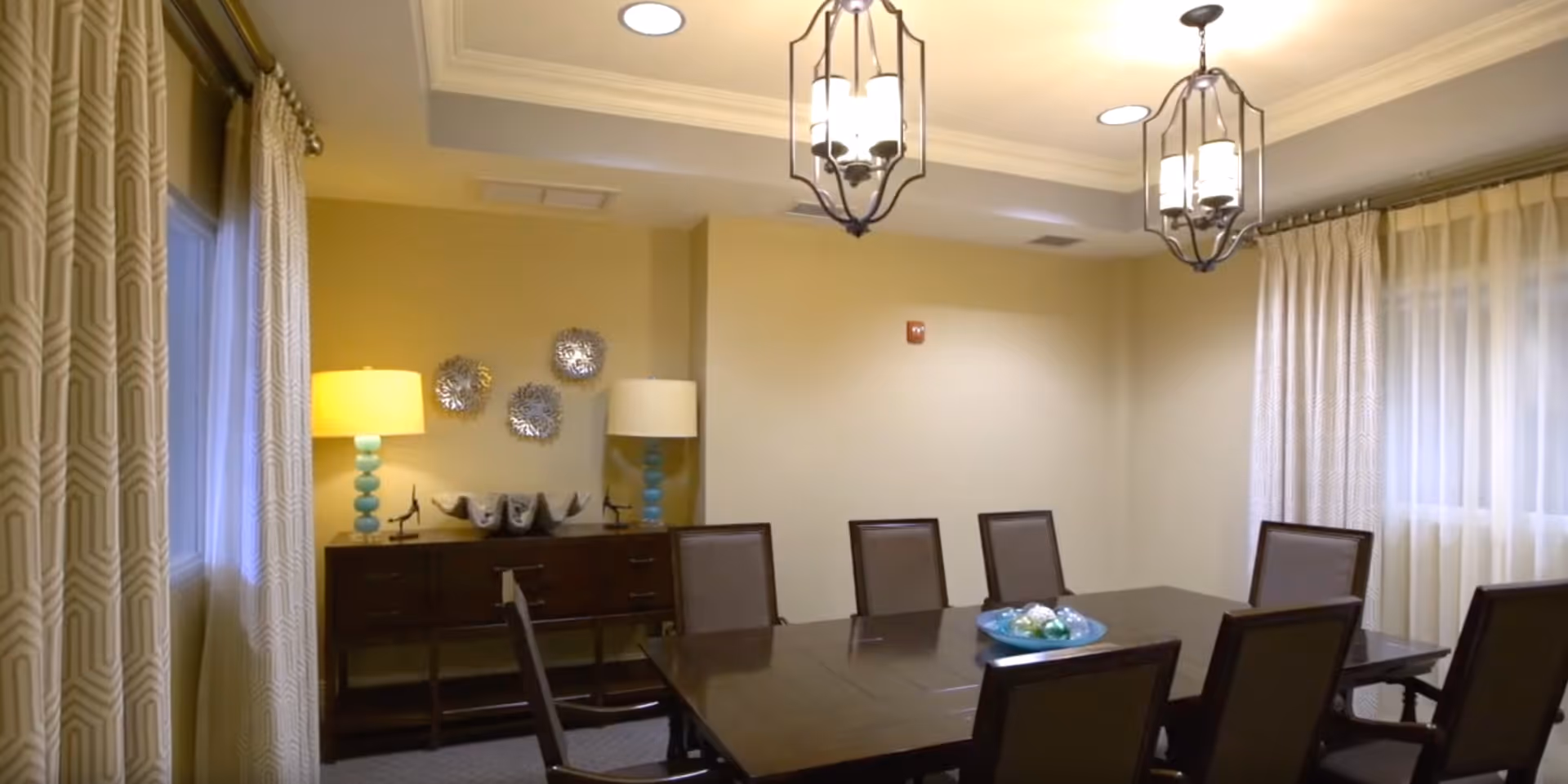 A well-lit dining room with a dark wooden dining table surrounded by eight chairs. The room features two large windows with patterned curtains, two decorative hanging light fixtures, and a sideboard with two lamps and decorative items on top. The walls are painted a soft beige color.