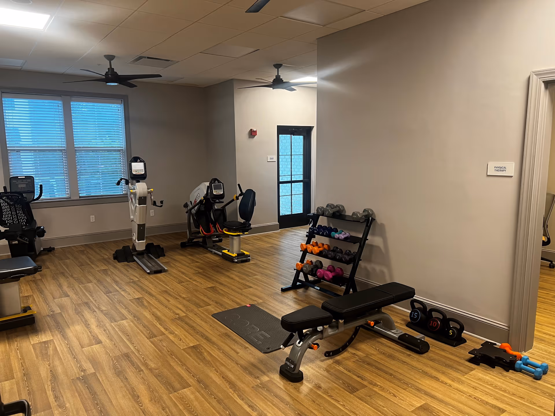 A bright exercise/physical therapy room with cardio machines, a weight bench, dumbbell rack, and wood-look flooring.
