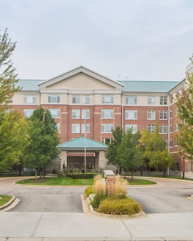 Front exterior view of a multi-story senior living facility building with a brick facade, green roof, and an entrance canopy. The driveway curves around landscaped greenery and trees are visible on both sides.