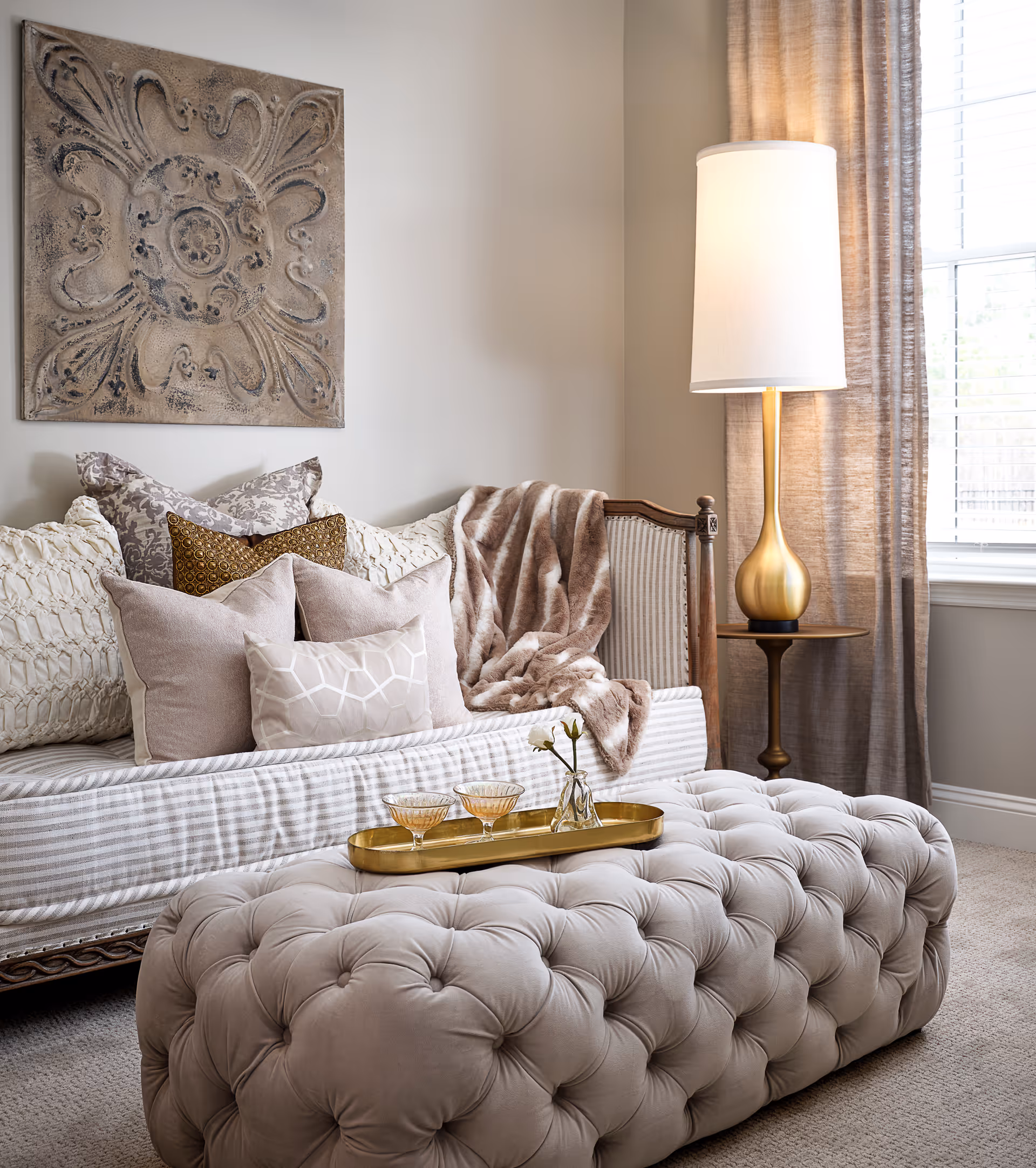A cozy living room corner featuring a tufted beige ottoman with a gold tray holding two glasses and a small vase with flowers. Behind it is a striped sofa adorned with multiple decorative pillows and a soft throw blanket. To the right, a tall gold lamp with a white shade stands next to a window with beige curtains.