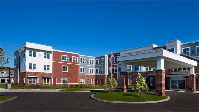 Exterior view of a modern three-story senior living facility building with a covered entrance, brick and siding facade, and a clear blue sky.