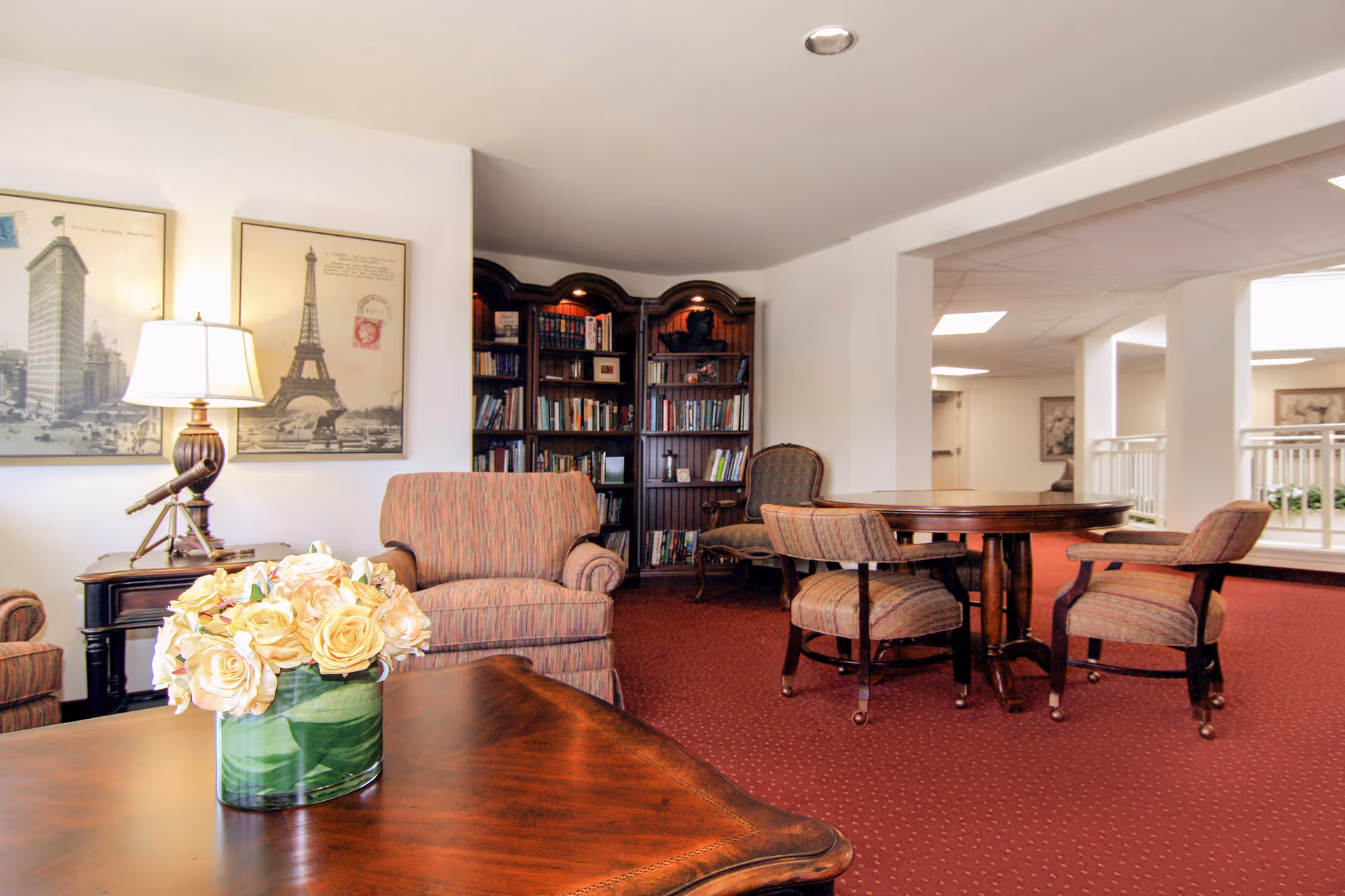 A cozy living room area with a wooden coffee table holding a vase of cream-colored roses, upholstered armchairs, a round wooden table with four chairs, a bookshelf filled with books, and framed pictures of the Eiffel Tower and a cityscape on the wall.
