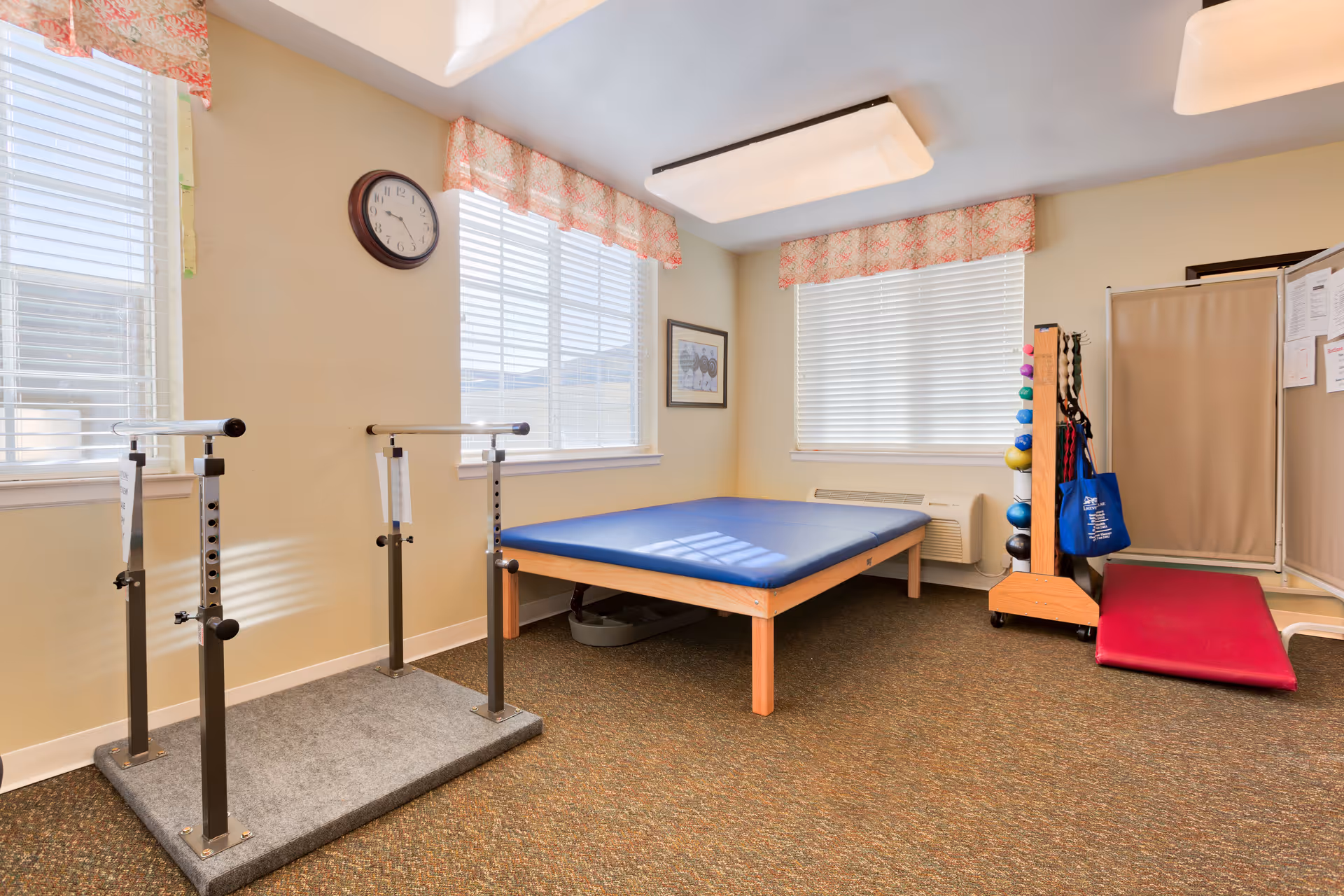 A small physical therapy or exercise room with a blue padded therapy table, parallel bars for walking support, a rack with colorful exercise balls and resistance bands, a red exercise mat, beige walls, two large windows with blinds and patterned valances, a wall clock, and overhead lighting.