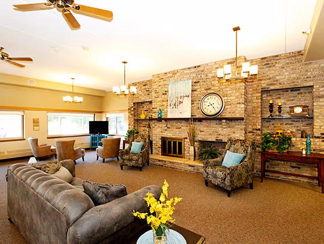 A spacious living room with a large gray sofa, two patterned armchairs with blue cushions, and a brick fireplace with a clock and artwork above it. The room has beige carpet, ceiling fans, and multiple light fixtures. There are windows letting in natural light and additional seating with chairs and a TV in the background.