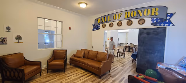 A seating area with two brown leather armchairs and a matching sofa arranged on a wooden floor. On the wall above the doorway is a decorative sign that reads 'WALL OF HONOR' with military emblems and a blue ribbon design. Through the doorway, a man is visible standing in a room with tables and chairs. The walls are light-colored, and there are framed tree artworks on the left wall.