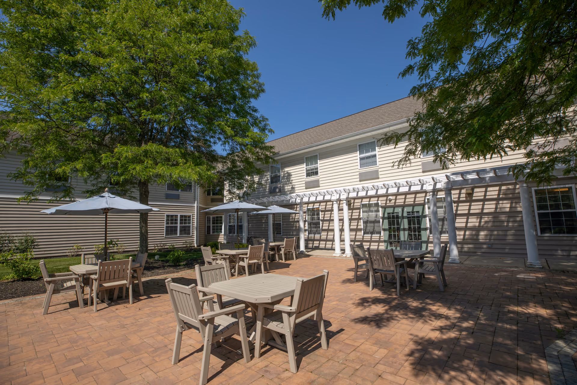 Outdoor patio area at Dresher Estates by Priority Life Care featuring multiple tables with chairs and umbrellas, surrounded by trees and the exterior of a two-story building under a clear blue sky.