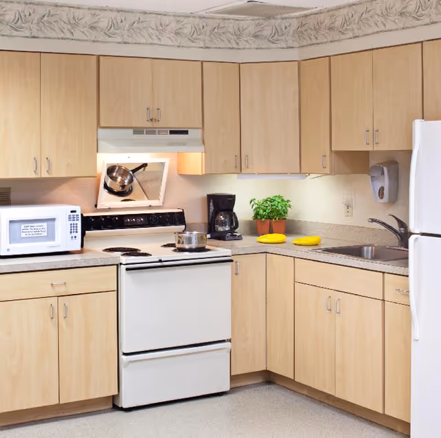 A clean kitchen with light wood cabinets, a white electric stove with a pot on it, a microwave, a coffee maker, a small potted plant, two yellow plates, a sink with a faucet, and a white refrigerator. The kitchen has a floral wallpaper border near the ceiling.