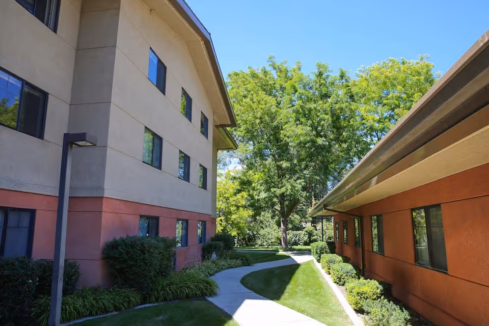 Outdoor view of a senior living facility showing two buildings with beige and reddish-brown walls, a curved concrete pathway between them, green bushes, grass, and trees under a clear blue sky.