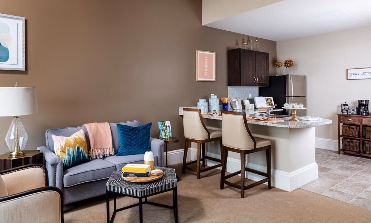 A cozy living area with a gray sofa adorned with colorful pillows and a pink throw blanket, a small side table with a lamp, and a patterned coffee table with books and decor. Adjacent to the living area is a kitchen with a granite countertop breakfast bar, two beige bar stools, dark wood cabinets, a stainless steel refrigerator, and a coffee station on a wooden cabinet.