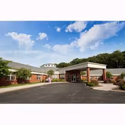 Exterior view of a single-story brick building with a covered entrance and a driveway, surrounded by greenery and trees under a partly cloudy blue sky.