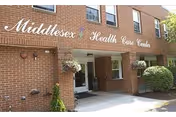 Exterior view of Middlesex Health Care Center showing a brick building entrance with the facility name displayed above the doorway. There are hanging flower baskets and some greenery near the entrance.