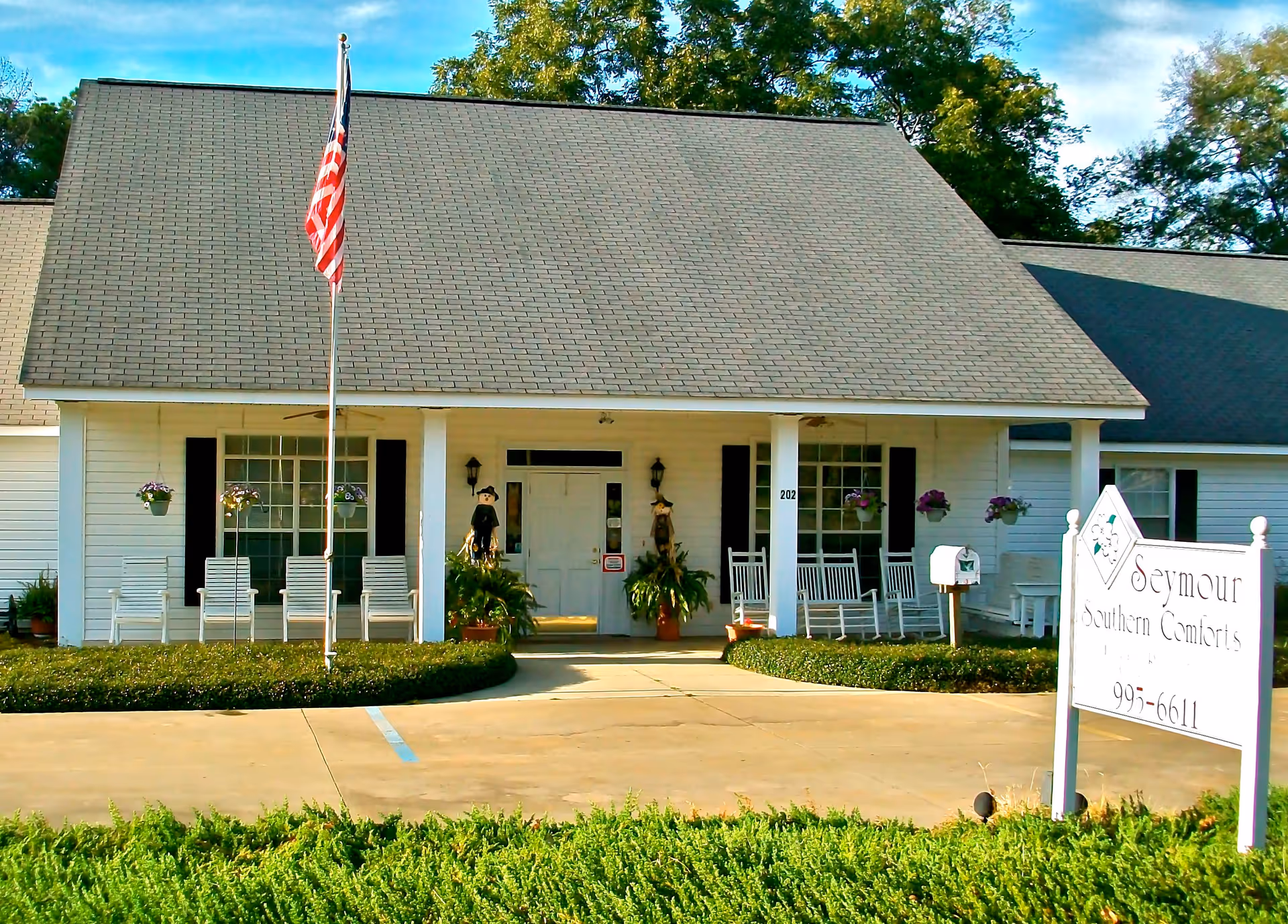 Front exterior view of a single-story building with a large sloped roof, white siding, and black shutters. There is a covered porch with white rocking chairs and hanging flower pots. An American flag is mounted on a pole in front of the building. A white sign near the entrance reads 'Seymour Southern Comforts'.