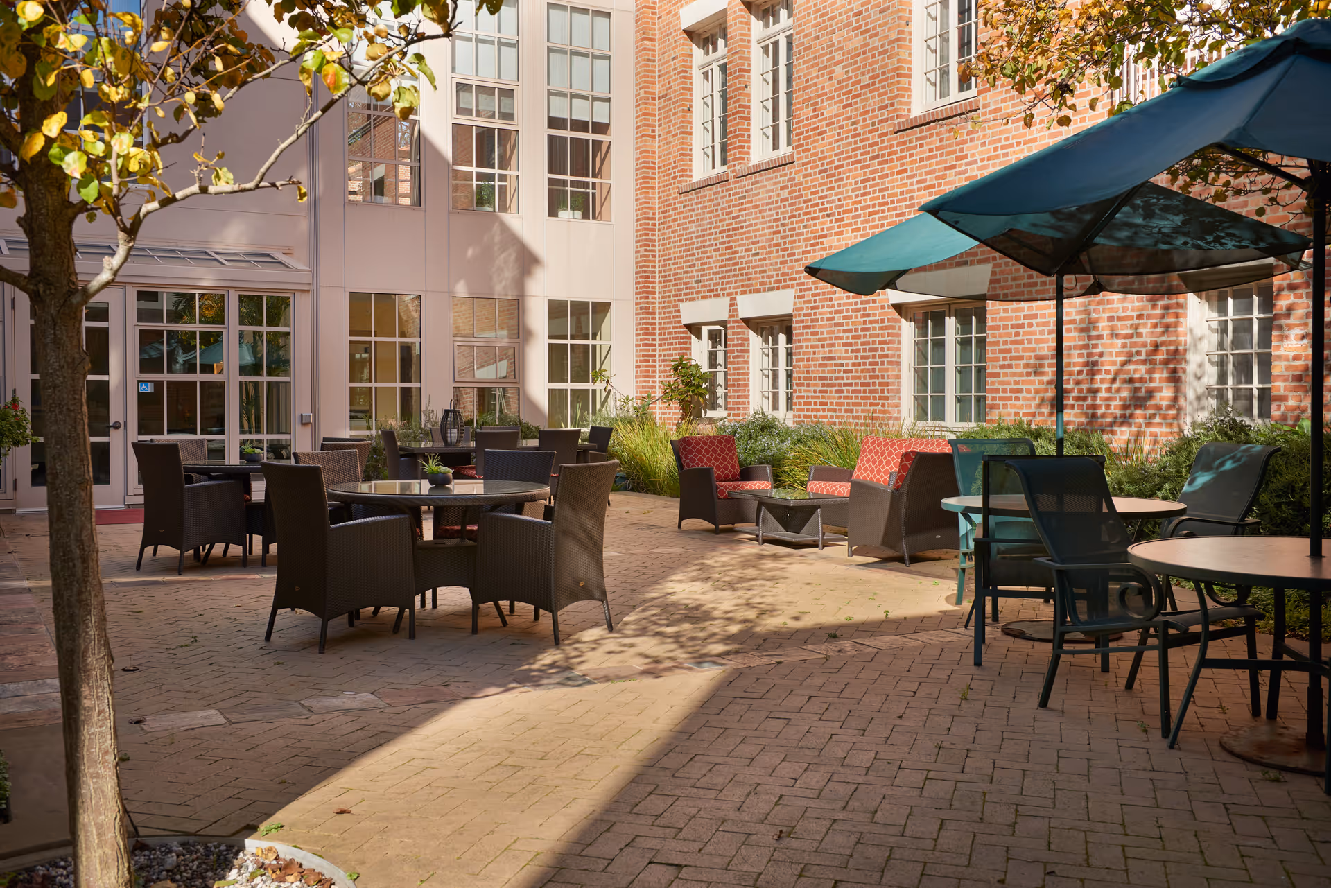 Outdoor patio area with multiple seating arrangements including round tables with chairs and cushioned lounge chairs. The patio is paved with bricks and surrounded by a building with large windows and red brick walls. There are green umbrellas providing shade over some tables and a tree with autumn leaves in the foreground.