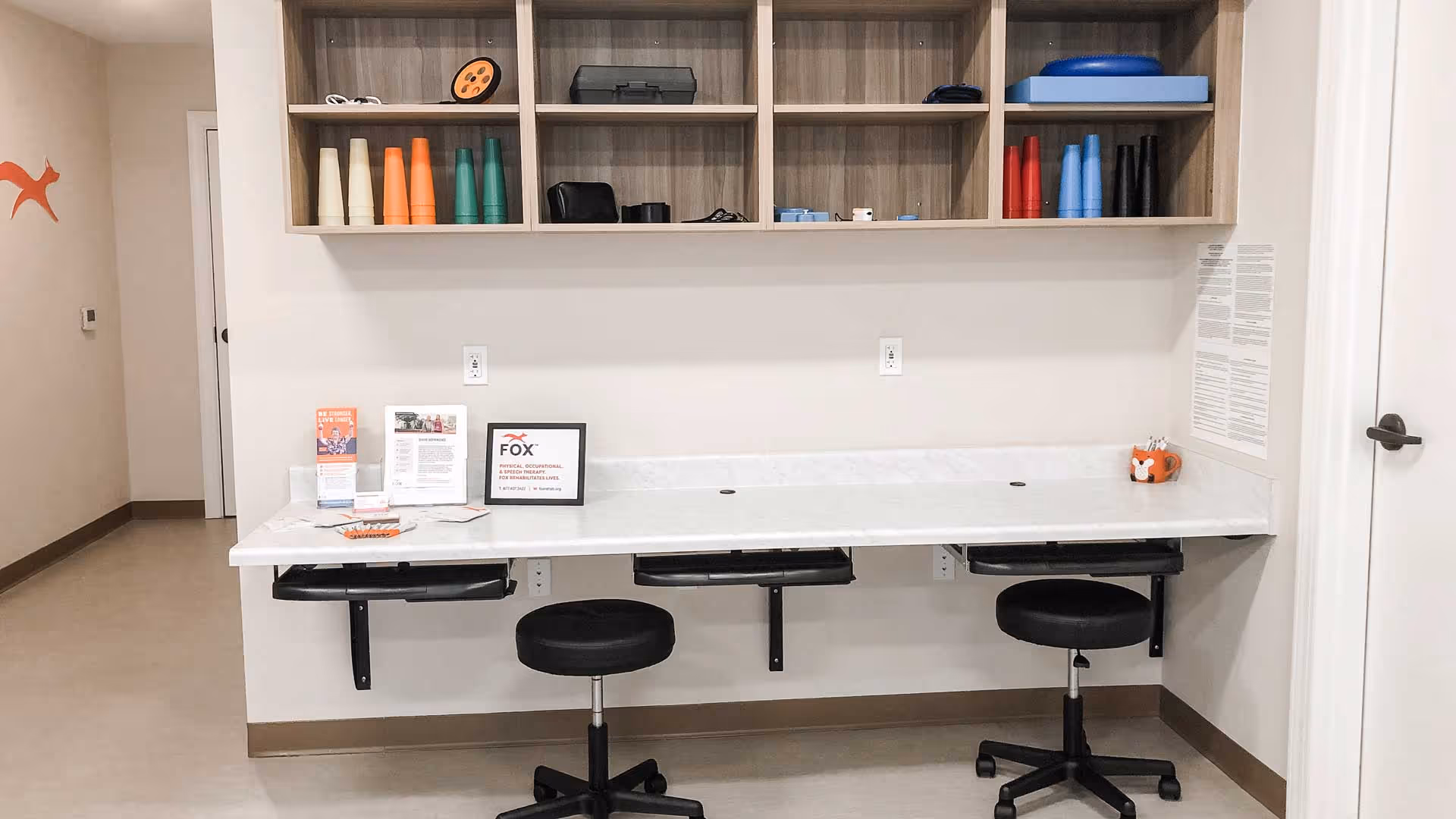 A clean, organized workspace with a long white countertop mounted to the wall and three black swivel stools underneath. Above the counter are wooden shelves holding various colored cones and small equipment. On the counter are informational pamphlets, a framed sign for FOX Physical, Occupational & Speech Therapy, and a small orange cup with pens. The room has light-colored walls and flooring, with a door on the right side and a small orange fox logo on the left wall.