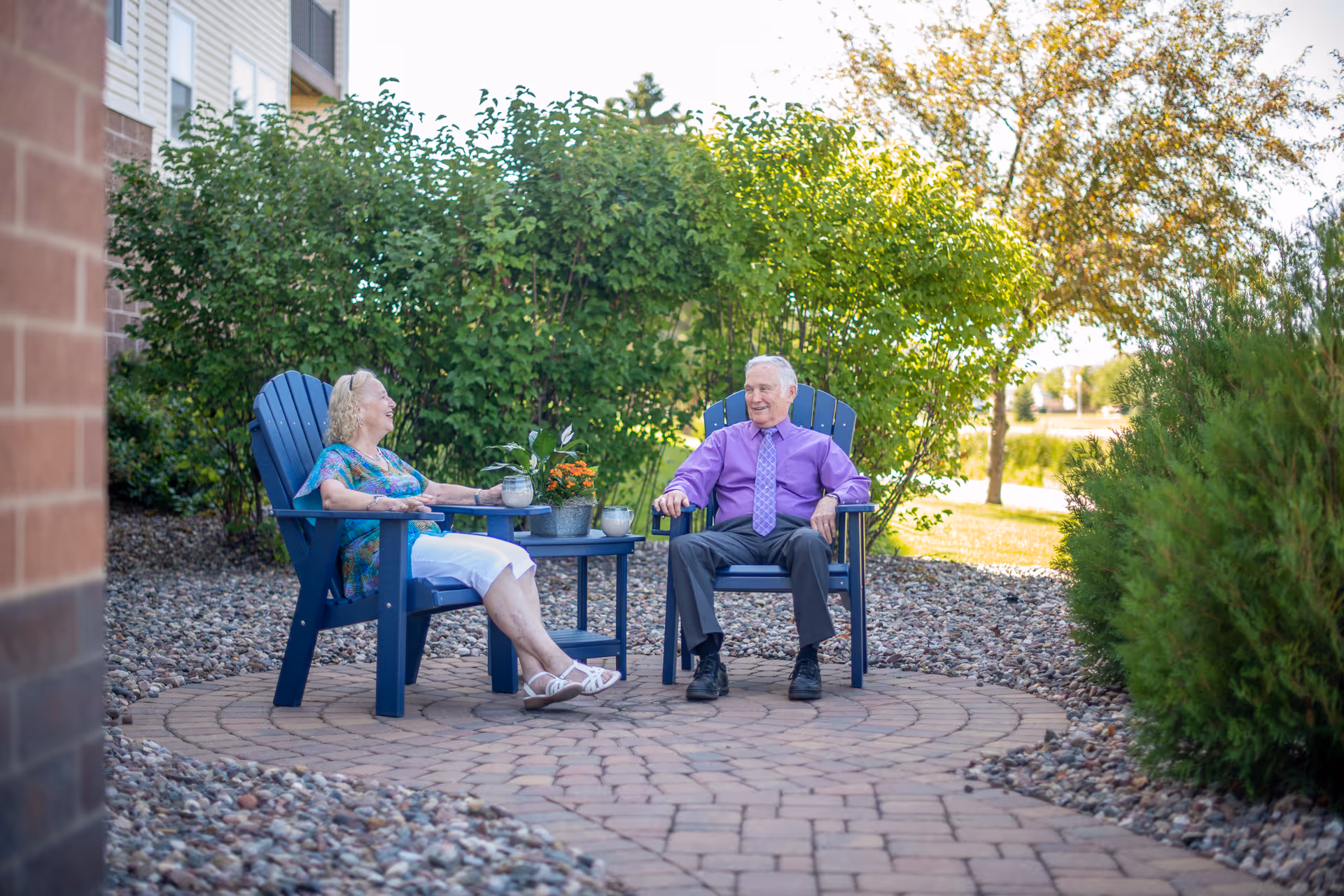 An elderly man and woman sitting outdoors on blue Adirondack chairs around a small table with potted plants, surrounded by greenery and a stone pathway at Shorewood Senior Campus.