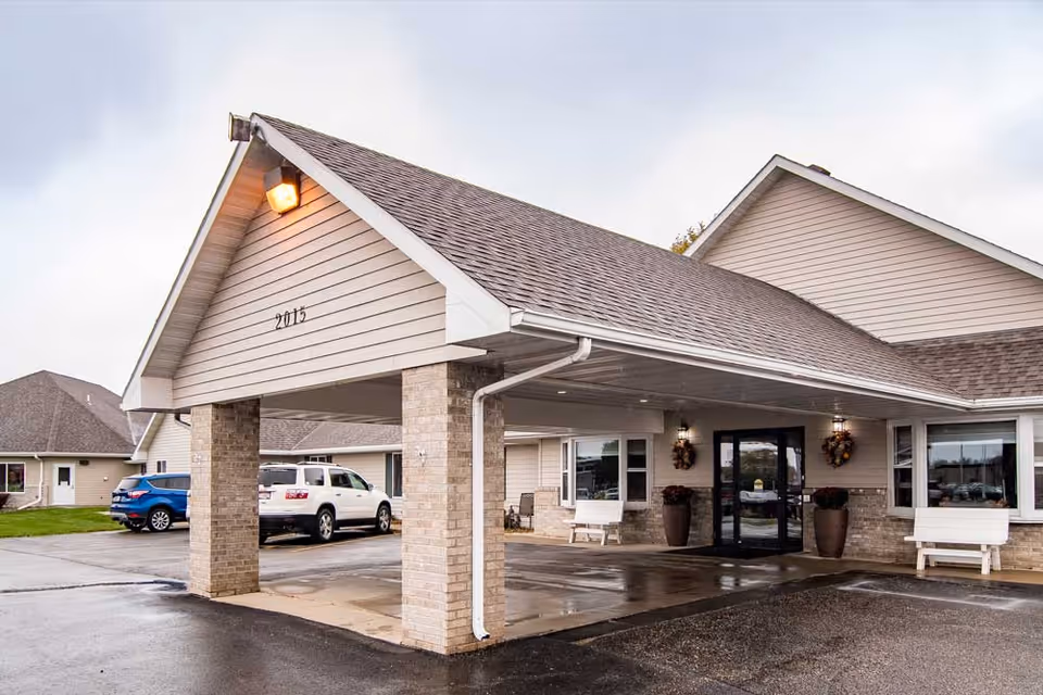 Exterior view of the entrance to Edgewood Brookings facility with a covered drop-off area supported by brick columns. There are two white benches and large potted plants near the glass double doors. Several cars are parked nearby on a wet driveway under an overcast sky.